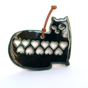 Puede incluir: Adorno de cerámica en forma de gato verde oscuro con recortes de corazón. El gato tiene un acabado brillante y un lazo de cuero marrón para colgar. El adorno está sobre un fondo blanco.