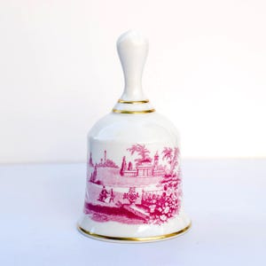 Puede incluir: Campana de cerámica blanca con un diseño de paisaje rosa y ribete dorado. La campana tiene un asa y una base acampanada. El diseño rosa representa un paisaje escénico con edificios, árboles y figuras. Un artículo decorativo.