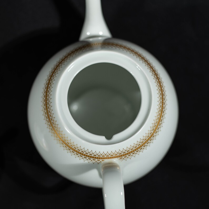 May include: White ceramic teapot with a gold decorative border around the rim. The teapot has a handle and a spout. The design is simple and elegant, suitable for serving tea. The teapot is set against a black background.