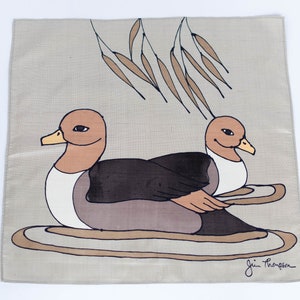 May include: A square, beige fabric with a hand-drawn illustration of two ducks. The ducks are brown, black, and white, with yellow beaks. Above the ducks are brown leaf-like shapes. The artist's signature is in the bottom right corner.