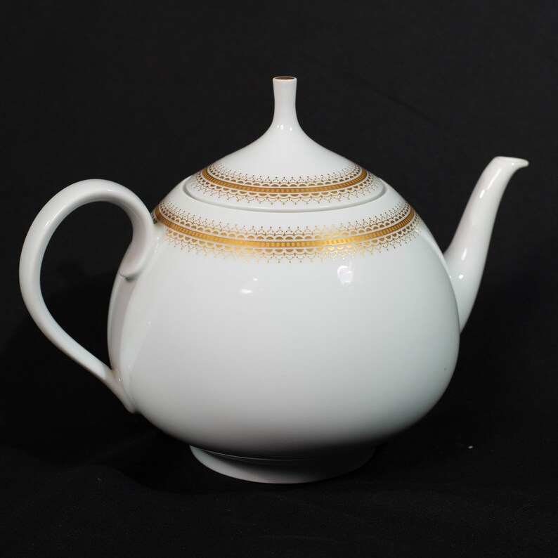 May include: White ceramic teapot with a rounded body, curved handle, and spout. The lid and body are adorned with a gold filigree pattern. The teapot sits on a black surface.