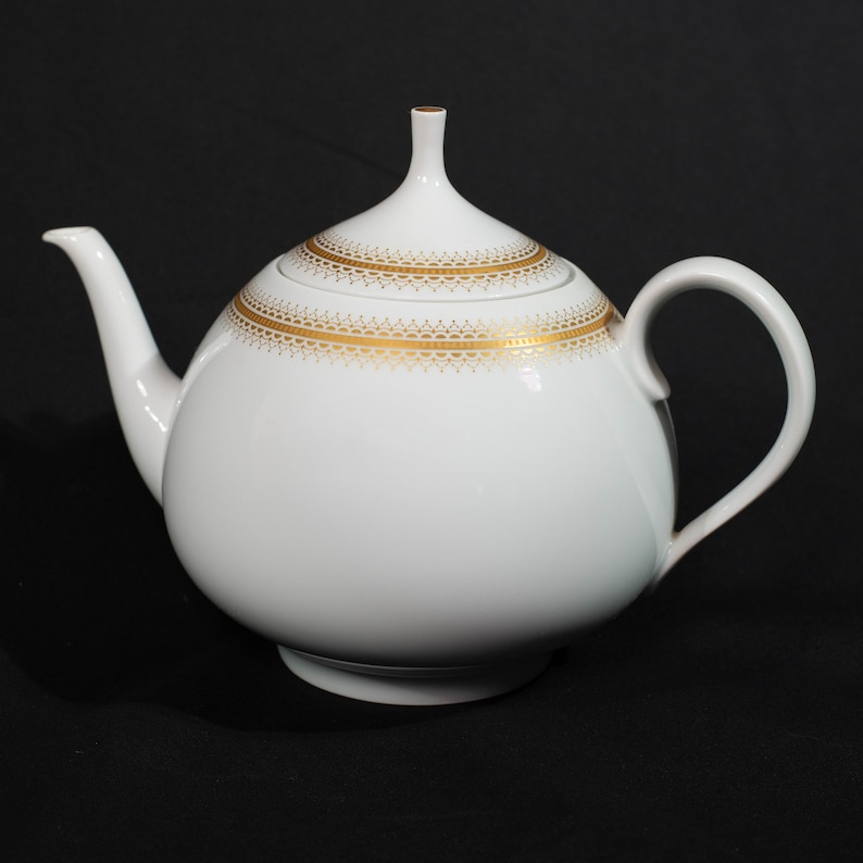 May include: White ceramic teapot with a rounded body, a curved spout, and a loop handle. The lid and body are adorned with gold filigree patterns. The teapot sits on a black surface.
