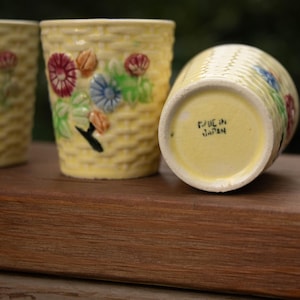 May include: A set of three yellow ceramic cups with a raised basket-weave texture and hand-painted floral motifs in red, blue, and green. The base of one cup is clearly marked with the text Made in Japan, indicating its origin and manufacturing.