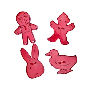 May include: A set of four red plastic cookie cutters featuring distinct shapes including a gingerbread person, a figure with a hat, a rabbit head with long ears, and a duck. Each cutter has a molded surface with raised details and a central handle.