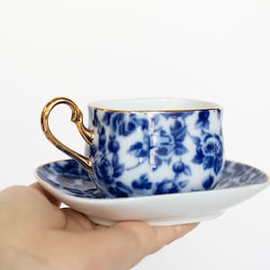 Puede incluir: Una taza de té y un platillo de porcelana blanca con un patrón floral azul y dorado. La taza tiene un borde dorado y un asa dorada.