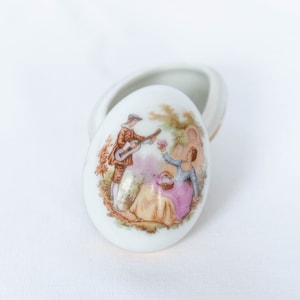 May include: A white porcelain egg-shaped trinket box with a lid. The lid features a painted scene of a couple in a garden setting. The man is playing a guitar and the woman is holding a basket of flowers.