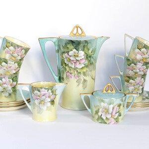 May include: A vintage porcelain tea set featuring a teapot, creamer, sugar bowl, and several teacups with saucers. The set is painted with a floral design in pastel colors, including pink, white, and green. The teapot is approximately 10 inches tall.