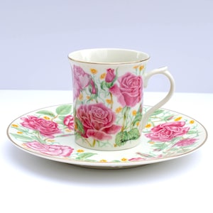 May include: A white porcelain teacup and saucer set with a floral design featuring pink roses and green leaves. The set has a gold trim around the edges.