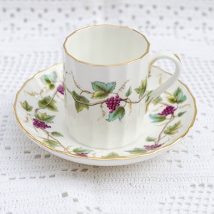 Puede incluir: Una taza de té y un platillo de porcelana blanca con un borde dorado y un diseño floral con hojas verdes y uvas moradas.