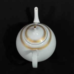 May include: White ceramic teapot with a gold filigree design around the lid. The teapot has a rounded body, a curved handle, and a long, slender spout. The lid has a small knob and a gold rim. The teapot is set against a black background.