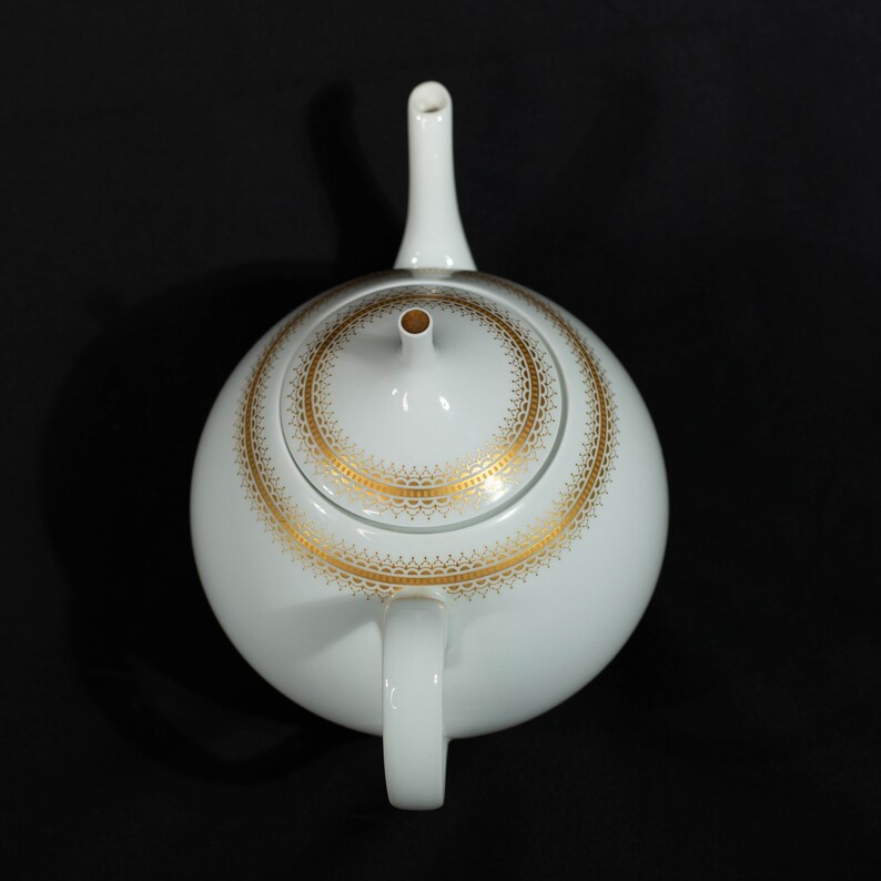 May include: White ceramic teapot with a gold filigree design around the lid. The teapot has a rounded body, a curved handle, and a long, slender spout. The lid has a small knob and a gold rim. The teapot is set against a black background.
