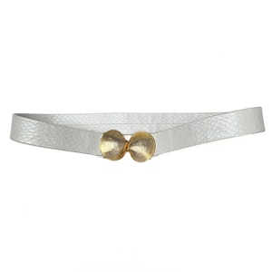 May include: A white belt featuring a textured snakeskin-like pattern across the strap. The center of the belt is secured with a metallic gold-toned buckle composed of two conjoined, ribbed, shell-shaped circular pieces that interlock together.