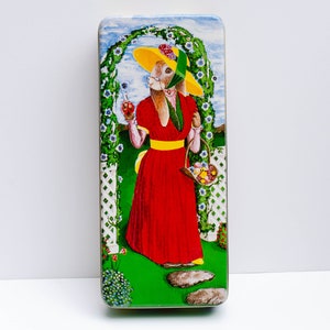 May include: A vintage tin box with a colorful illustration of a rabbit wearing a yellow hat and red dress, holding a basket of fruit and flowers. The rabbit is standing in a garden setting with a white trellis and green foliage.