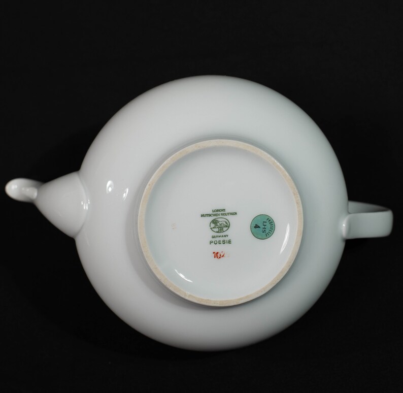 May include: A white porcelain teapot with a rounded body, spout, and handle. The bottom of the teapot features the text "Poesie" and the brand name "Lorenz Hutschenreuther Germany". A green sticker with the number "4" is also visible.