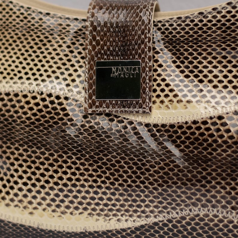 May include: Close-up of a handbag with a snakeskin pattern in shades of brown and beige. The bag features a rectangular flap with the text "MONICA BY MAGLI" on a silver plate. The bag's texture is visible.