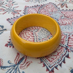 May include: A solid, smooth, mustard yellow bangle bracelet with a thick, rounded profile and a high-gloss finish. The circular accessory features a simple, continuous band design with no additional embellishments, patterns, or hardware.