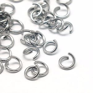 May include: A selection of small, silver jump rings scattered on a white background. The rings are open, with a small gap for attaching to other jewellery components. The metal has a shiny, reflective appearance.