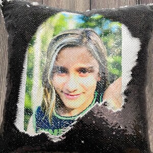 May include: A black and silver sequin pillow with a photo of a young person smiling. The photo is printed on a white background and shows the person's face and shoulders.