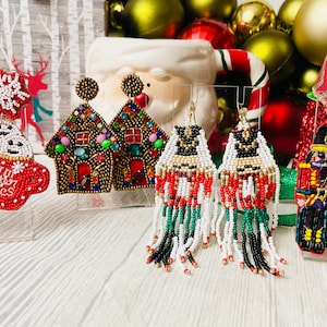 Può includere: Assortimento di orecchini con perline a tema natalizio. Gli orecchini presentano una casetta di pan di zenzero, una tazza con il testo "Warm Winter Wishes", uno schiaccianoci e un fiocco di neve.