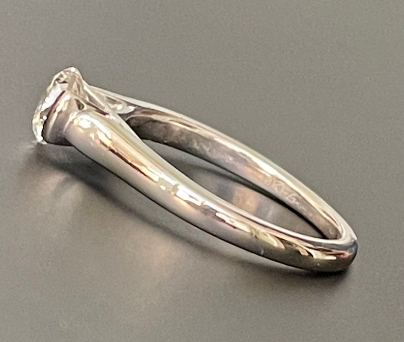 K&ouml;nnte beinhalten: Ein silberner Verlobungsring mit einem einzelnen runden Diamanten, der in einer Krappenfassung sitzt. Die Band ist d&uuml;nn und hat ein leicht geschwungenes Design.