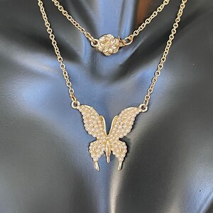 Könnte beinhalten: Eine goldene Kettenkette mit einem zarten Schmetterlingsanhänger. Der Schmetterling ist aus Gold und hat eine perlmuttartige Oberfläche. Die Kette ist mit einer zweiten Kette geschichtet, die einen kleinen Blumenanhänger hat.