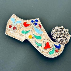 Könnte beinhalten: Eine silberne Brosche in Schuhform mit bunten Emaildetails. Der Schuh ist mit floralen Mustern in Rot, Blau, Grün und Gelb verziert. Die Ferse des Schuhs ist ein Cluster aus silbernen Perlen.