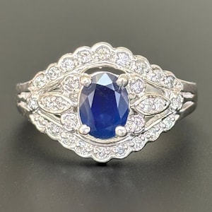 Könnte beinhalten: Ein silberner Ring mit einem großen ovalen blauen Saphir, der von kleineren Diamanten umgeben ist. Der Ring hat ein zartes, filigranes Design mit einem floralen Motiv.