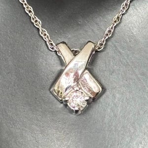 Könnte beinhalten: Eine silberfarbene Halskette mit einem Anhänger in X-Form. Der Anhänger hat einen kleinen, runden, klaren Edelstein in der Mitte. Die Kette ist filigran und ebenfalls silberfarben, was einen klassischen und eleganten Look erzeugt.