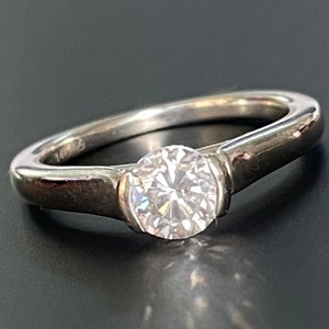 K&ouml;nnte beinhalten: Ein silberner Ring mit einem einzigen, runden, klaren Diamanten in einer Zargenfassung. Die Band ist schlicht und elegant.