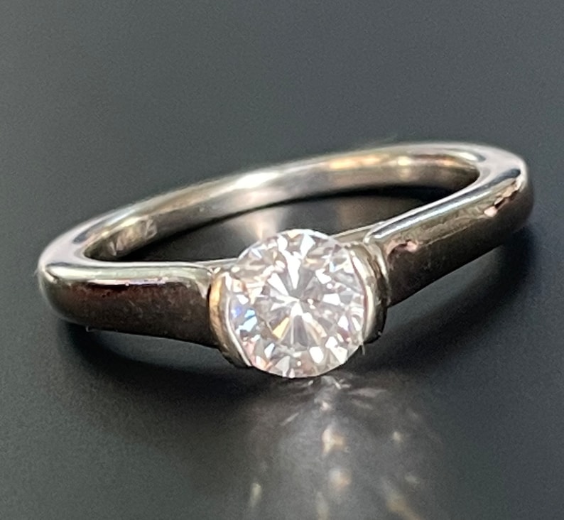 K&ouml;nnte beinhalten: Ein silberner Ring mit einem einzigen, runden, klaren Diamanten in einer Zargenfassung. Die Band ist schlicht und elegant.