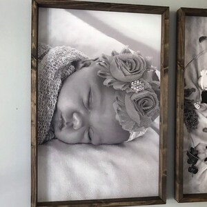 May include: A black and white photo of a sleeping baby wearing a flower crown. The photo is framed in a dark wood frame.