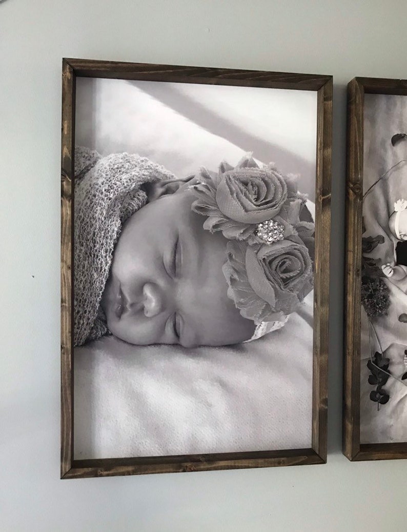 May include: A black and white photo of a sleeping baby wearing a flower crown. The photo is framed in a dark wood frame.