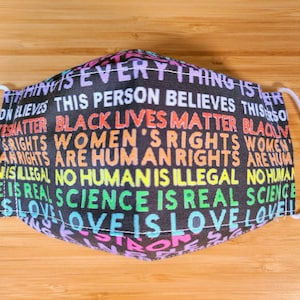 May include: Black cloth face mask with white elastic ear loops. The mask has a repeating pattern of colorful text that reads "Everything is...