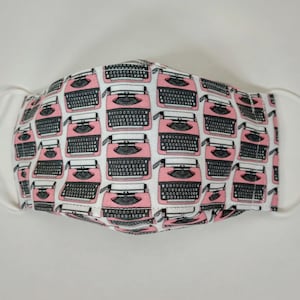 May include: A white face mask with a pink and black typewriter pattern. The mask has two white elastic straps.