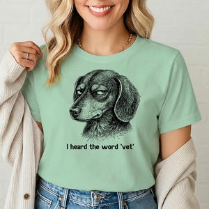 May include: A pale green t-shirt with a black and white drawing of a dachshund dog with a dubious expression. The text "I heard the word 'vet'" is printed below the dog.