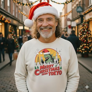 May include: A person wearing a cream-coloured sweatshirt featuring a Godzilla graphic in a Santa hat over a Tokyo skyline. The text reads "Merry Christmas from Tokyo" in red and white. The person is also wearing a Santa hat.