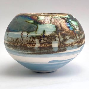 May include: A round ceramic bowl featuring a smooth, light blue and white base with horizontal stripes. The upper portion displays a textured, metallic glaze in shades of bronze, dark green, and iridescent blue with a crackled, weathered finish.