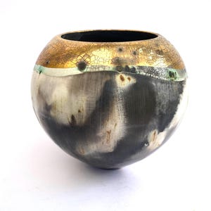 Puede incluir: Un jarrón redondo de cerámica con un esmalte negro y blanco. La parte superior del jarrón está decorada con una banda de oro y verde.