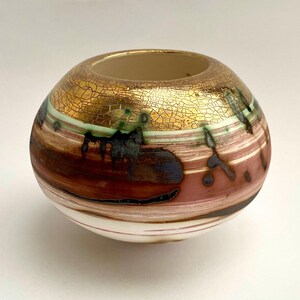 Könnte beinhalten: Eine dekorative Keramikvase mit runder Form und breiter Öffnung. Der obere Bereich ist goldfarben mit einer craquelé-Textur. Der mittlere Bereich weist horizontale Streifen in Braun-, Rosa- und Grüntönen auf. Der Boden ist weiß.