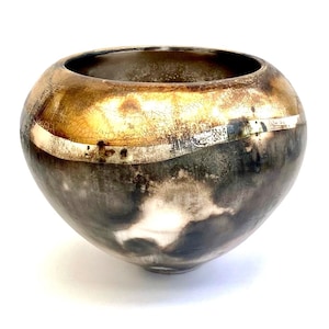 Puede incluir: Un cuenco de cerámica con una base ancha y redondeada y un borde ligeramente acampanado. El cuenco está decorado con una banda de esmalte metálico dorado alrededor de la parte superior, con un efecto craquelado. El resto del cuenco es de color marrón oscuro con toques de blanco y gris.