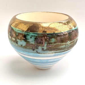 May include: A ceramic bowl with a wide, rounded top and a tapered base. The top is gold with a crackled texture, transitioning to a turquoise and brown band. The lower portion features horizontal blue and white stripes.
