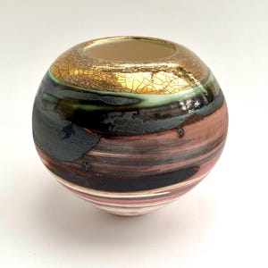 Peut inclure: Un vase sphérique en céramique avec un dessus doré craquelé et une ouverture circulaire. Le vase présente des bandes horizontales de couleurs, notamment des nuances de marron, noir, vert et rose. L'intérieur est beige clair.