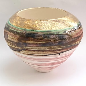 May include: A handcrafted ceramic bowl with a wide opening and a rounded shape. The top section features a gold, crackled glaze, followed by a green band, and then layers of brown, pink, and white horizontal stripes. The interior is a light beige color.