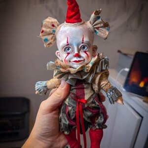 May include: A handmade clown doll with a white face, red accents, and blue eyes. The doll wears a red and black patterned outfit with a ruffled collar and a red cone hat. The doll's shoes are red with gold accents.