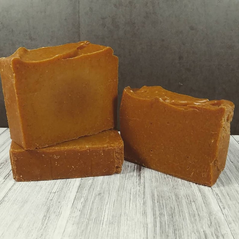 Puede incluir: Tres barras rectangulares de jab&oacute;n artesanal. El jab&oacute;n es de color marr&oacute;n claro y tiene una superficie texturizada. Las barras est&aacute;n apiladas y dispuestas sobre una superficie de madera blanca y gris. El fondo es gris oscuro.