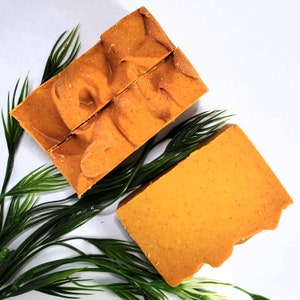 Puede incluir: Dos barras de jab&oacute;n hechas a mano con una superficie texturizada de color amarillo anaranjado. Una barra est&aacute; cortada en dos trozos. Ramitas verdes y frondosas est&aacute;n dispuestas alrededor de las barras de jab&oacute;n sobre un fondo blanco. El jab&oacute;n tiene un aspecto natural y org&aacute;nico.