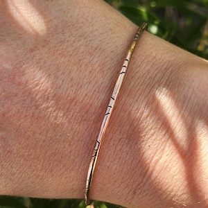 May include: A simple gold bangle bracelet with a textured design. The bracelet is worn on a person's wrist.