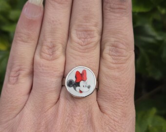 May include: A silver ring featuring a circular design with a cartoon image of Minnie Mouse. The ring has a white background with Minnie's face, a red bow, and black ears. The ring is on a finger.