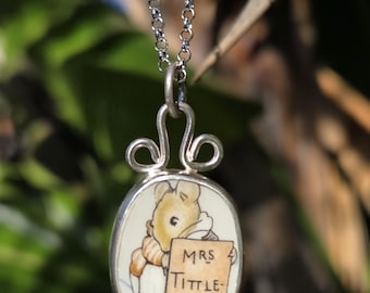 May include: Silver-toned pendant necklace featuring a small, oval-shaped charm with a cartoon illustration of a mouse holding a sign that reads "MRS TITTLE-MOUSE". The charm is suspended from a delicate chain.