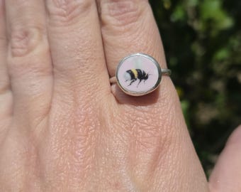 May include: A silver ring with a round, pink face featuring a detailed illustration of a bumblebee. The bee is black and yellow, set against a soft pink background. The ring is shown on a person's finger.